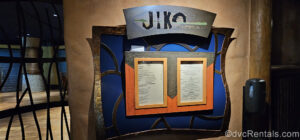 The blue, orange, and grey metal sign for Jiko – The Cooking Place is seen outside the restaurant at Jambo House. The menu is also displayed in smaller orange frames.