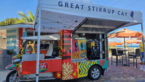 A multicolored mini truck with drink dispensers called the “Tiki Zoom Bar” is seen under a white canopy reading “Great Stirrup Cay.” Behind the truck there is another bar, as well as tables with orange umbrellas, and the ocean can be seen in the distance.