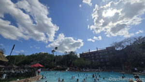 Guests swim in the pool at Disney’s Animal Kingdom Villas Jambo House under a sunny, blue sky. The dark brown resort buildings can be seen in the background