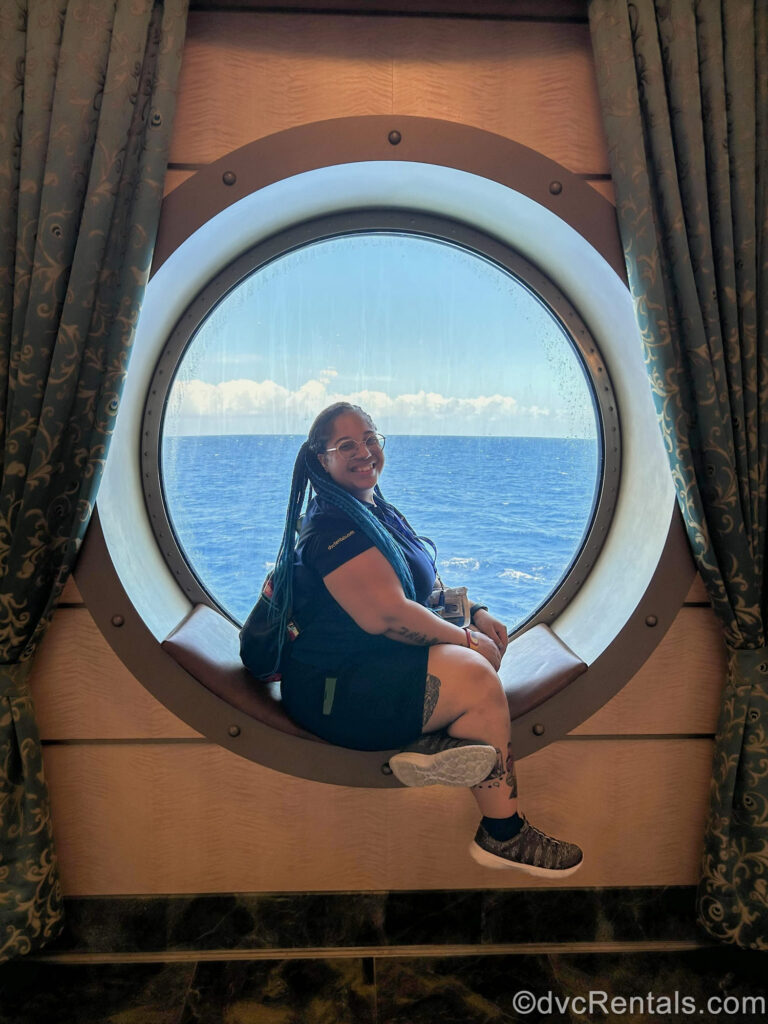 David’s Vacation Club Rentals team member Mya poses with a smile while sitting on a porthole window bench aboard the Disney Fantasy.