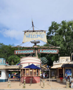The entrance to Disney’s Typhoon Lagoon Water Park at Walt Disney World is seen under a blue sky. A tall shipwreck sail reads “Welcome!” in blue lettering, while two wooden planks reading “Typhoon Lagoon” hang below. Guests are seen entering the park under the signs.