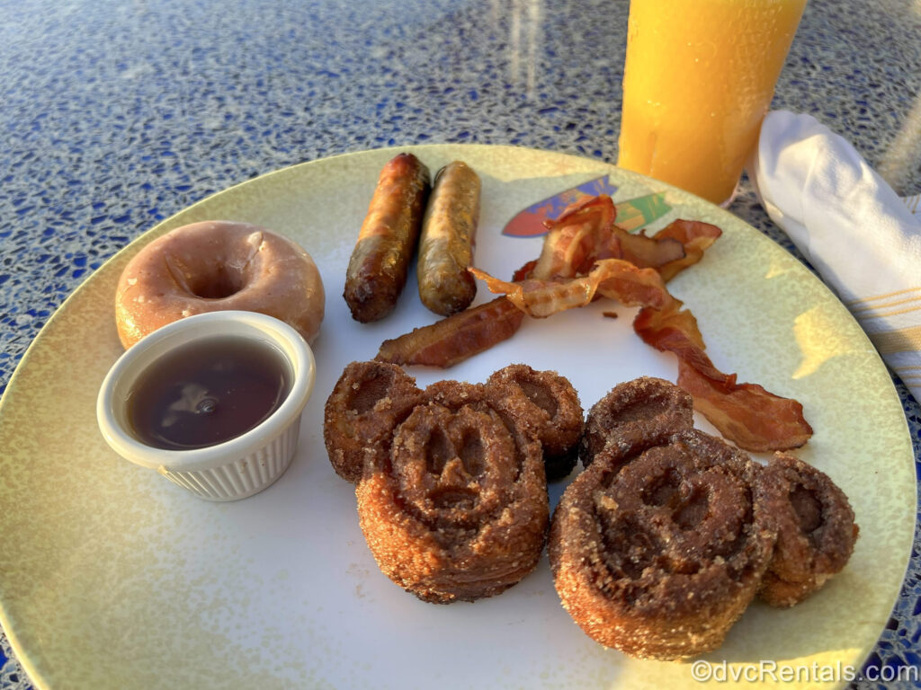 2 Churro Mickey Waffles covered in cinnamon sugar are seen on a white and yellow plate along with bacon, sausages, a donut, and a small bowl of maple syrup.