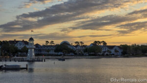 The sunrise makes for a beautiful orange, yellow, and blue sky over Disney’s Beach Club Villas. A boat can be seen gliding over Crescent Lake in front of the resort, and the Spaceship Earth ball at EPCOT is visible in the distance behind.