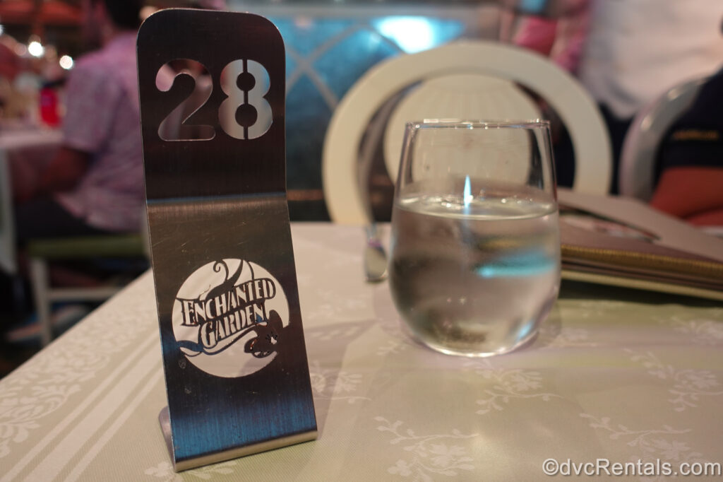 A silver table number marker with the dining room’s name cut into it “Enchanted Garden” sits atop a white tabletop in front of a glass of water.