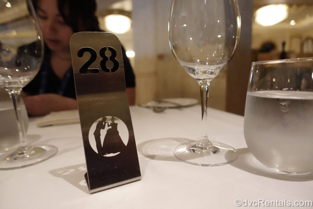 A Cinderella-inspired silver table number marker sits atop a white tablecloth in front of glass wine glasses and cups in the Royal Court dining room on the Disney Fantasy.