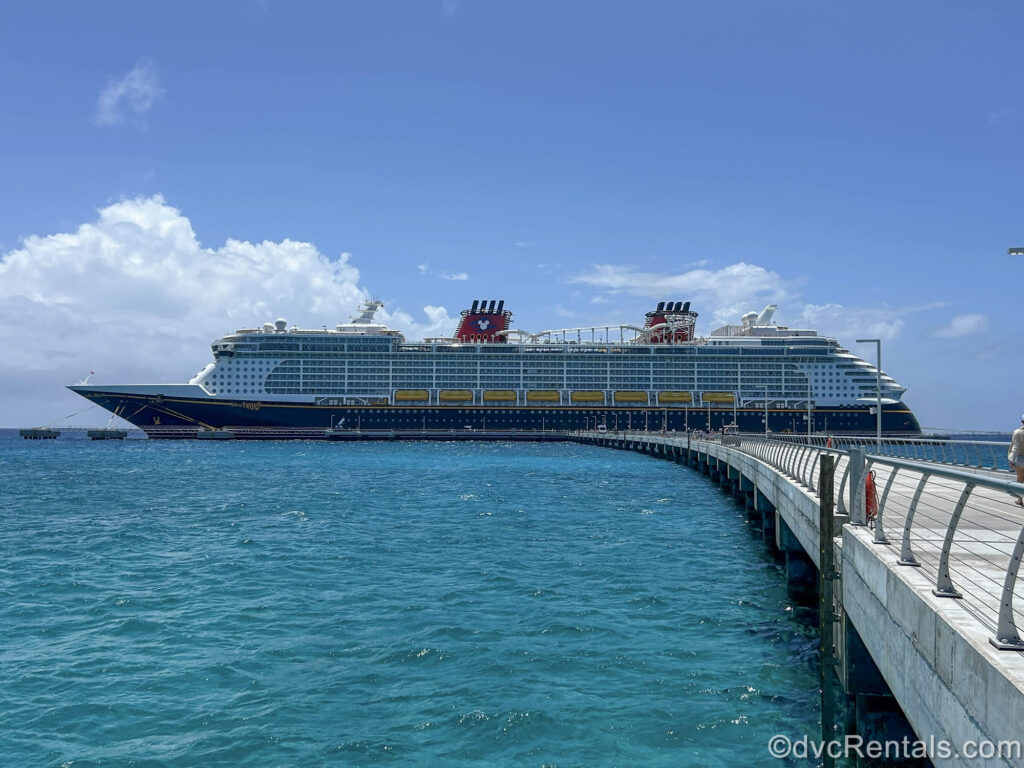 The Disney Fantasy cruise ship is seen floating on the water under a sunny blue sky from across the pier at Disney Lookout Cay at Lighthouse Point.