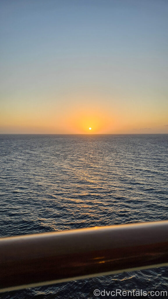 An orange and yellow sunrise alights the morning sky in the horizon over the ocean as seen from the Verandah of a Deluxe Oceanview Stateroom with Verandah on the Disney Fantasy.