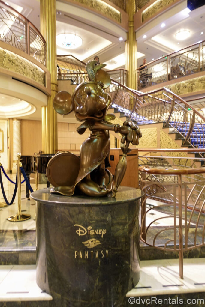 A bronze statue of Minnie Mouse with an umbrella, the atrium icon on the Disney Fantasy, is seen atop a marble pedestal reading “Disney Fantasy” in gold lettering with the Disney Cruise Line logo.