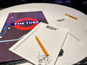 A trivia answers page and yellow pencils as well as a drinks menu sit on a white circular table in The Tube activity venue onboard the Disney Fantasy.