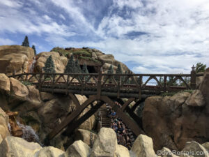 A train car full of guests speeds over the wood and steel tracks through the brown rocks of the Seven Dwarfs Mine Train under a blue sky full of fluffy white clouds.