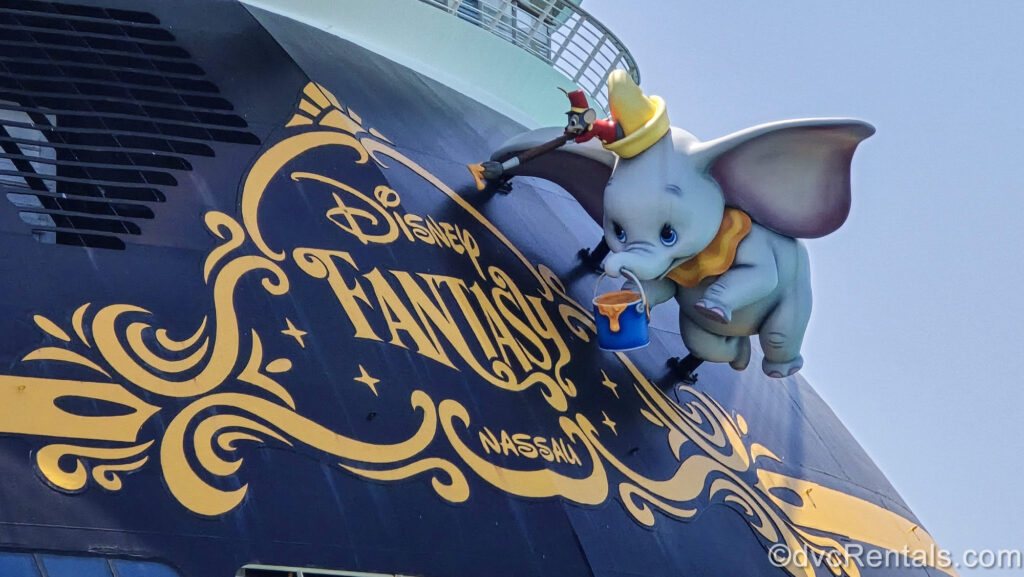 Large figures of Dumbo and Timothy Mouse are seen pretending to paint the exterior of the Disney Fantasy cruise ship. In yellow writing, the words “Disney Fantasy, Nassau” are painted on the ship.