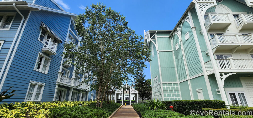 Building exteriors of Disney’s Beach Club Villas are seen under a blue sky with a walkway between two buildings. The building on the left is blue and white and the DVC villa building on the right is pastel green and white.