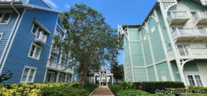 Building exteriors of Disney’s Beach Club Villas are seen under a blue sky with a walkway between two buildings. The building on the left is blue and white and the DVC villa building on the right is pastel green and white.