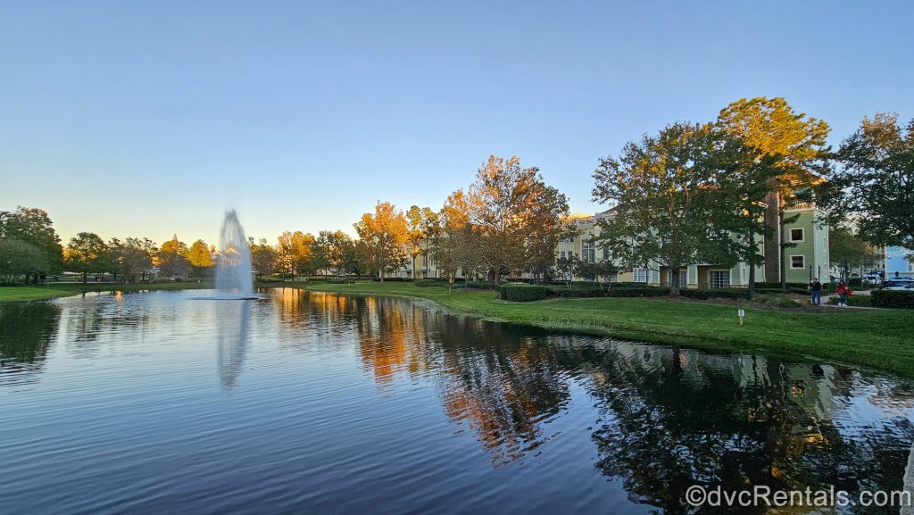 Golden Hour sunshine glows on the trees, water, fountain, lawns, and pastel buildings of Disney’s Saratoga Springs Resort & Spa under a dusky sky