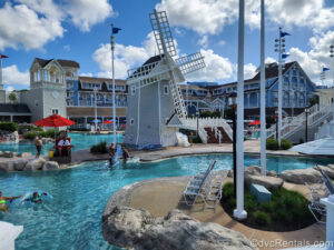 Guests enjoy the different pool areas at Stormalong Bay at Disney’s Beach Club Villas under a blue sky as the blue and white resort buildings are seen in the background.