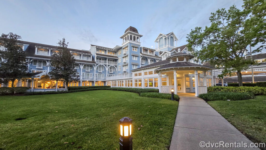 A green lawn and paved pathway lead to the entry into Disney’s Beach Club Villas, as the dawn sky brightens over the blue and white resort building.