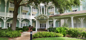 A walkway surrounding by green foliage leads to the mint green and white building that houses the DVC rooms at Disney’s Beach Club Villas. In front of the entry door is a statue of the Little Mermaid sitting on a brown rock with a sign reading “Disney Vacation Club” on it.