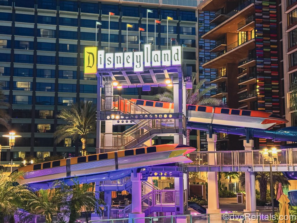 Two Monorail decorations and the retro Disneyland sign are seen overlooking the waterslide featured at the Villas at Disneyland Hotel at night as the lights glow over the pool area.
