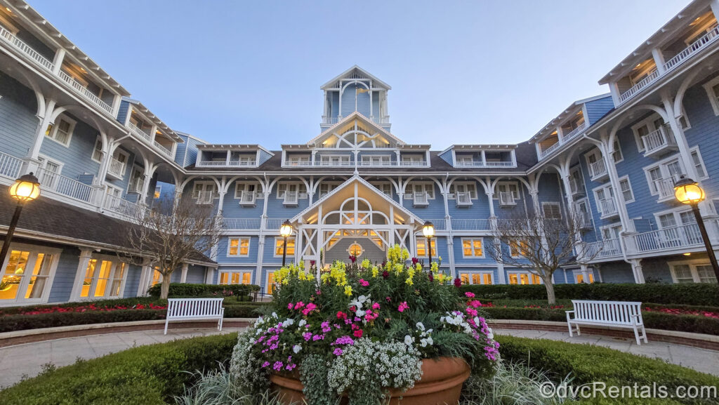 The blue and white exterior of Disney’s Beach Club Villas is seen as the sun begins rising under an early-dawn sky. The lampposts are still lit and there is a large planter with yellow, white, and pink flowers in front of the entrance doors.