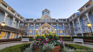 The blue and white exterior of Disney’s Beach Club Villas is seen as the sun begins rising under an early-dawn sky. The lampposts are still lit and there is a large planter with yellow, white, and pink flowers in front of the entrance doors.