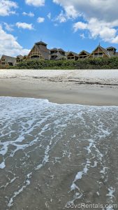Ocean waves wash up on the sandy beachfront as the exterior of Disney’s Vero Beach Resort is seen in the background under a blue sky.