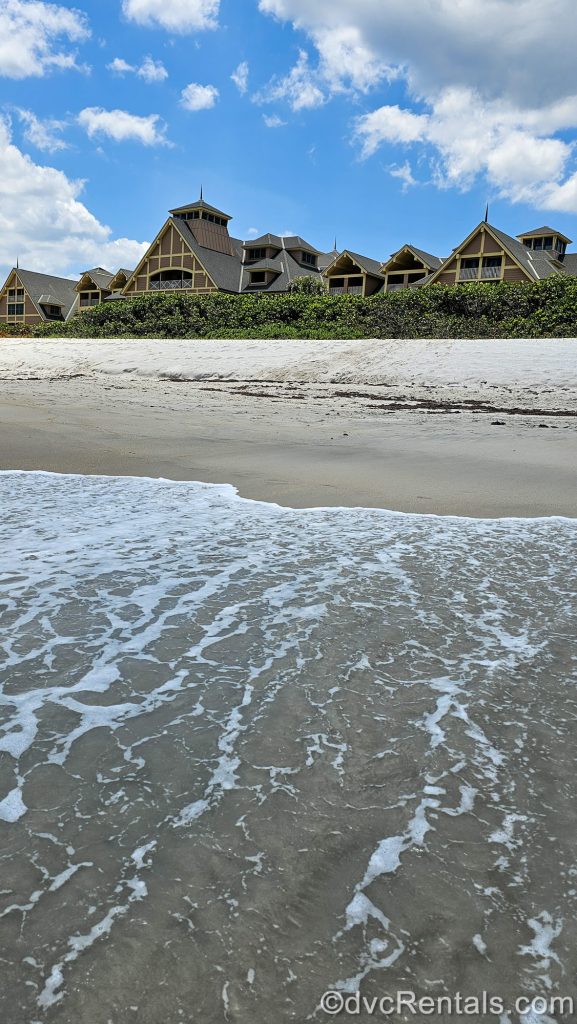 Ocean waves wash up on the sandy beachfront as the exterior of Disney’s Vero Beach Resort is seen in the background under a blue sky.