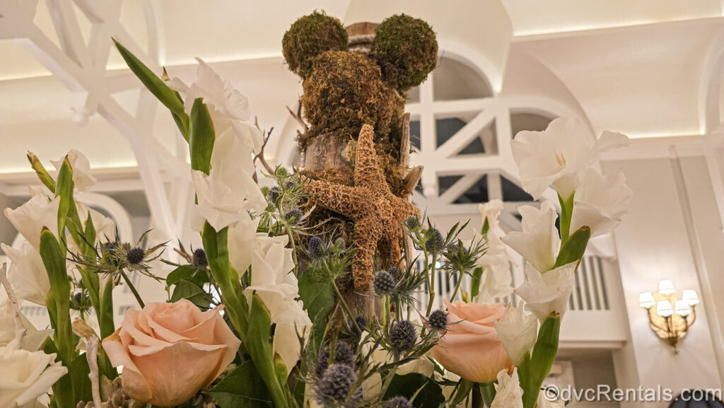 A Mickey Mouse head shaped out of green moss and foliage sits atop a floral display in the lobby of Disney’s Beach Club Villas. There is also a starfish, with pink roses, and other white flowers in the arrangement.