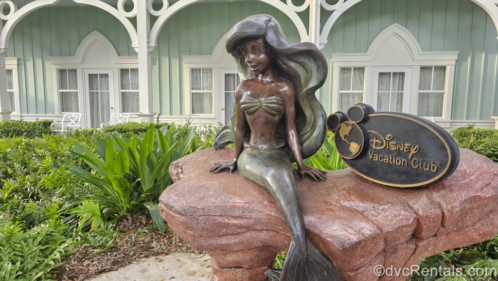 A statue of the Little Mermaid sits atop a stone outside the pastel green building next to a sign reading “Disney Vacation Club” at Disney’s Beach Club Villas