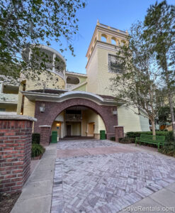 The pastel yellow, green and red brick entry into a building in the Grandstand section of Disney’s Saratoga Springs Resort & Spa is seen under a blue sky. There are green trees on either side of the entry as well as a green bench.