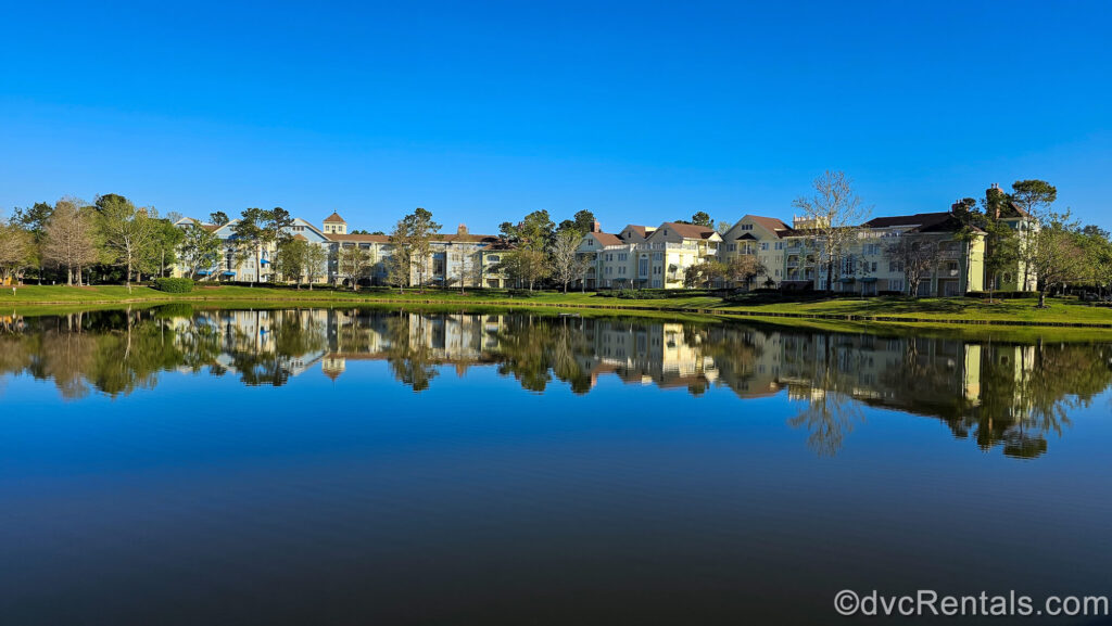 The pastel yellow, blue, and green buildings of Disney’s Saratoga Springs Resort & Spa are seen on green landscaping as its exterior is reflected in the body of water on the resort grounds under a blue sky.