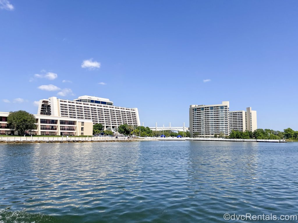 The white exterior buildings of Disney’s Contemporary Resort and Bay Lake Tower are seen from the waters of Bay Lake under a blue sky.