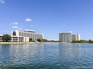 The white exterior buildings of Disney’s Contemporary Resort and Bay Lake Tower are seen from the waters of Bay Lake under a blue sky.