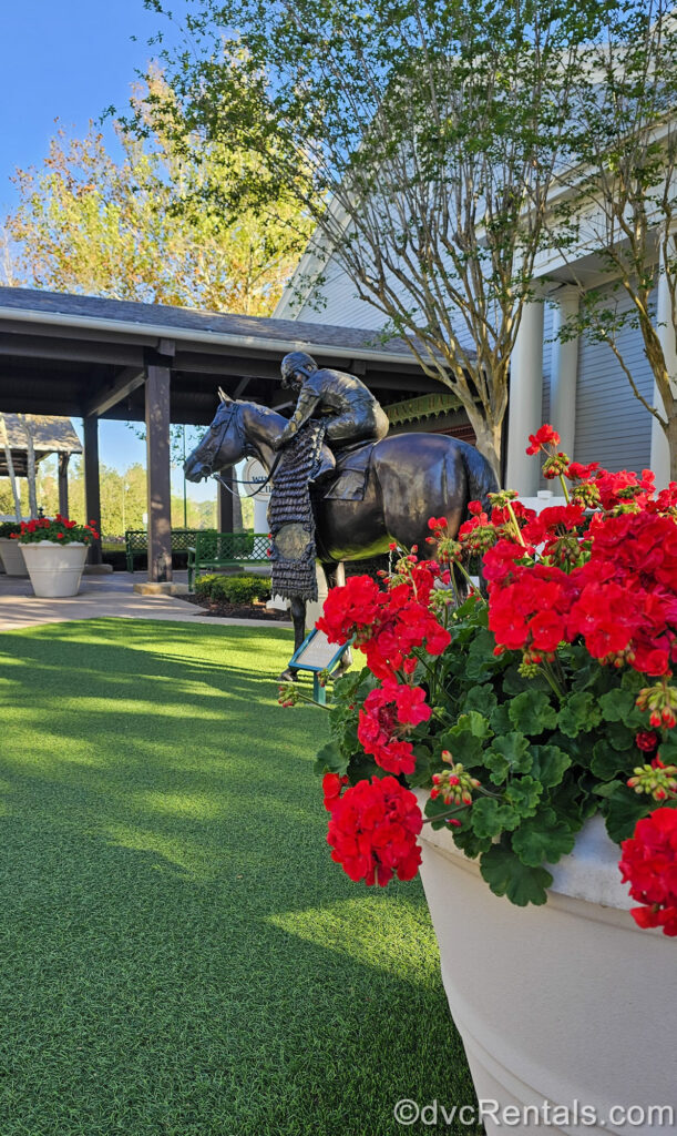 A planter full of red flowers is seen in front of the equestrian statue with horse and jockey outside the Carriage House at Disney’s Saratoga Springs Resort & Spa under a sunny sky.