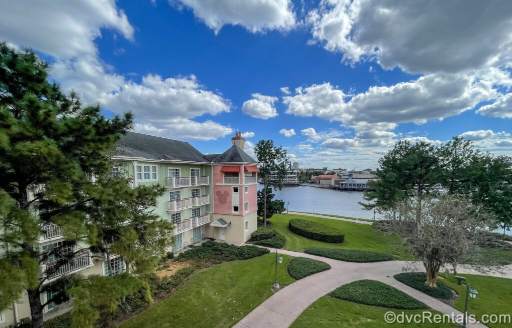 The pastel green, pink, and beige buildings of Disney’s Saratoga Springs Resort & Spa are seen under a blue sky. The resort’s green lawns are also seen with Disney Springs visible across the water in the distance.
