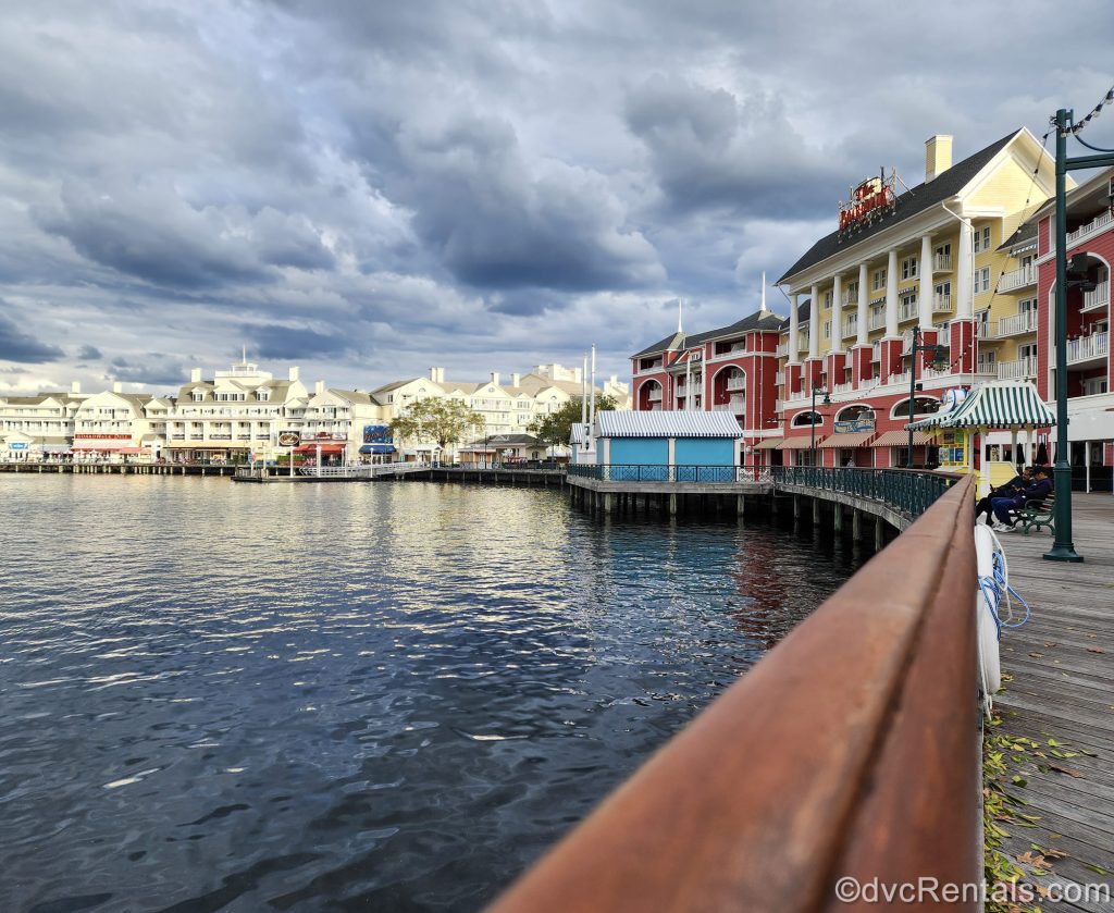 The pastel buildings of Disney’s BoardWalk Villas are seen along the waters of Crescent Lake and the BoardWalk itself under a cloudy sky.