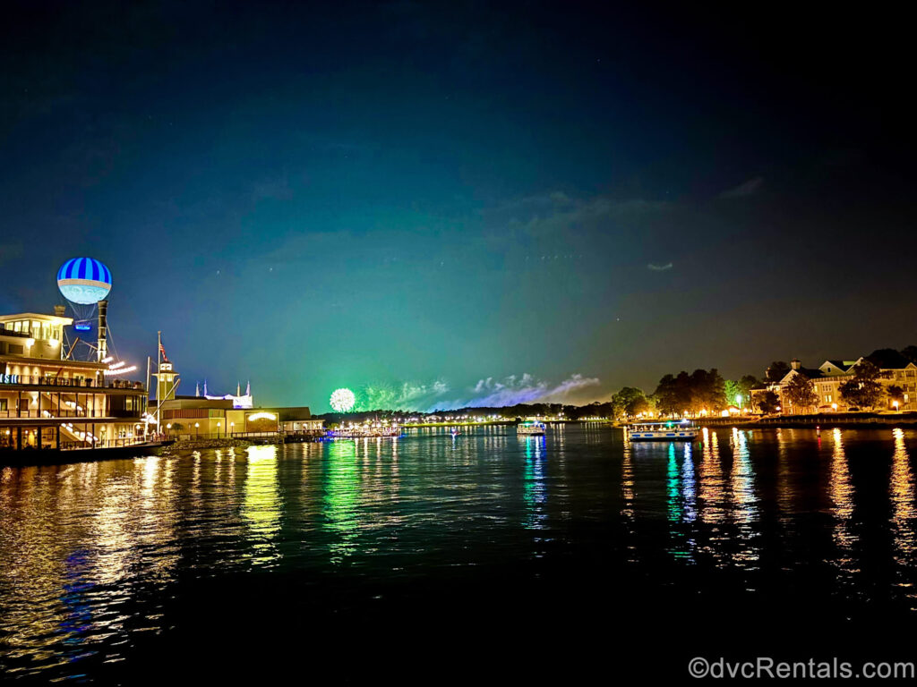 The lights of Disney Springs sparkle on the left as the warm glow of Disney’s Saratoga Springs Resort & Spa light up the right side of the photo across the water. In the distance of the night’s sky, theme park fireworks of green and blue are seen.