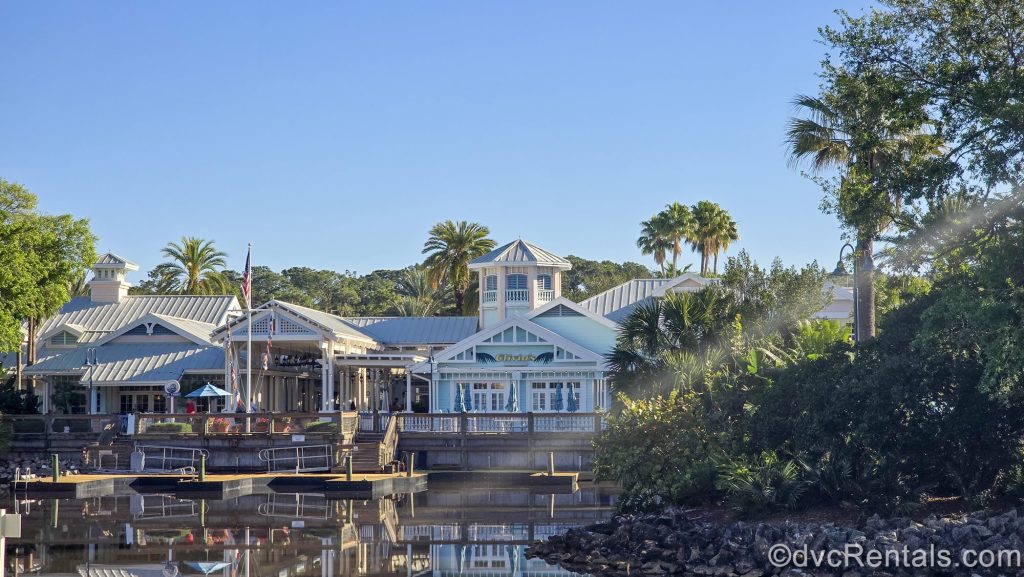 The pastel green and white exterior of Hospitality House and Olivia’s Café are seen under a sunny sky at Disney’s Old Key West Resort, with the calm river and wooden boat docks in front.