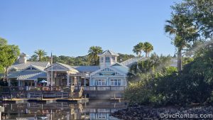The pastel green and white exterior of Hospitality House and Olivia’s Café are seen under a sunny sky at Disney’s Old Key West Resort, with the calm river and wooden boat docks in front.