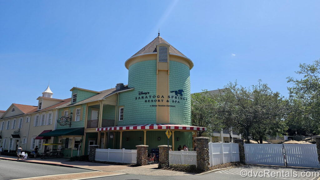 Soft green and yellow resort buildings housing amenities are seen under a sunny sky. The circular tallest portion of the corner building has the resort’s horse racing logo on it, as well as “Disney’s Saratoga Springs Resort & Spa, A Disney Vacation Club Resort” written in black lettering.