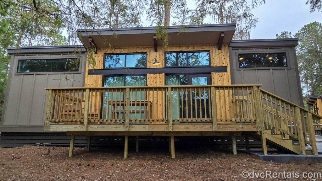 The brown and wooden exterior of a One Bedroom Cabin at Disney’s Fort Wilderness Resort is seen. There is a wooden staircase leading up to it and a deck with seating.
