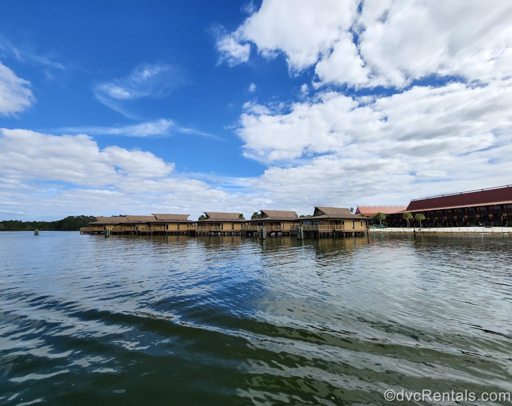 The exterior of a Longhouse at Disney’s Polynesian Villas and the over-water Bungalows are seen from across the Seven Seas Lagoon under a blue sky.