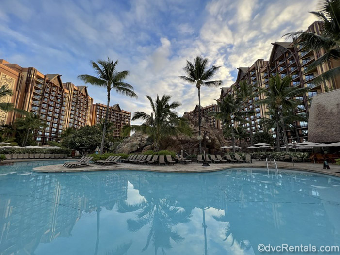 The exterior of Aulani, A Disney Resort & Spa is seen under a blue, morning sky with an empty pool, loungers, and palm tress in front of it.
