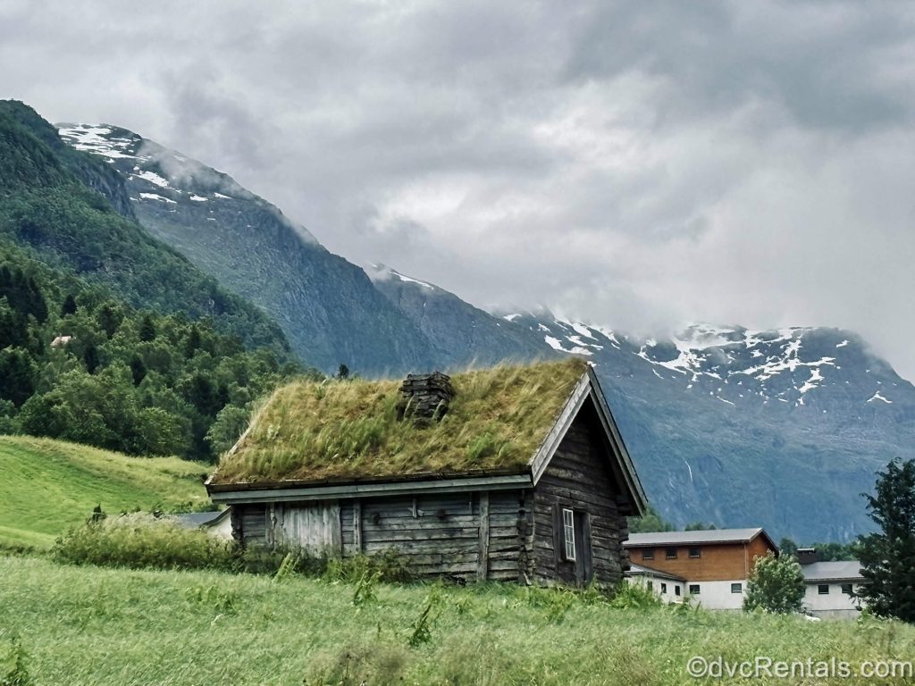 A wooden building with a green turf roof sits under a cloudy sky with snow-capped mountains and forests in the background in Olden, Norway.