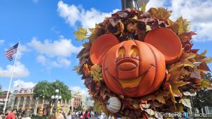 An orange Mickey-shaped pumpkin surrounded by a wreath of leaves hangs on a light post at the Magic Kingdom under a blue sky. Main Street U.S.A. and other Halloween decorations can be seen in the background.