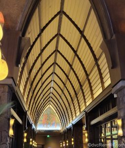 The lobby of Aulani is seen with a tall arched ceiling of yellow and dark wood. At the end there is an art piece depicting Hawaiians hunting for fish in the water.