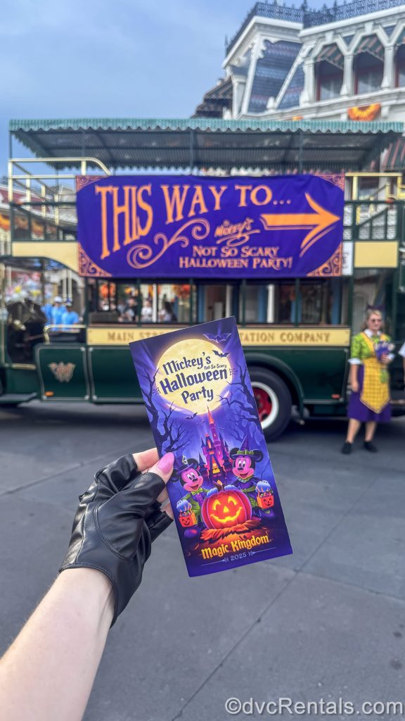 A hand holds a guide map for Mickey’s Not-So-Scary Halloween Party at Magic Kingdom. In the background there is an omnibus decorated for the event entrance.