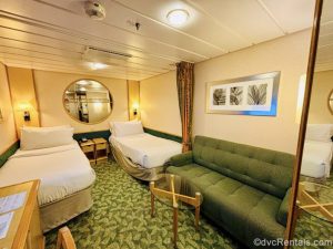 An Interior Stateroom with two twin beds, a large circular mirror, a green sofa, glass coffee table, and vanity with chair. The walls are cream colored with green patterned carpeting.