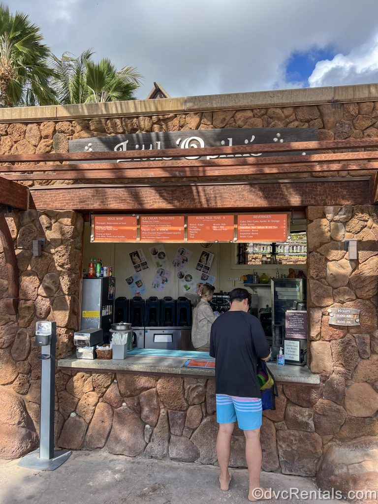 A man stands at the Little ‘Opihi’s snack stand as a Cast Member behind the counter prepares an order. The stand has a brown rock façade and an orange menu board.