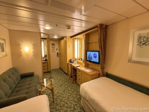 The living space of an Interior Stateroom onboard Independence of the Seas. Part of the two twin beds can be seen, as well as the TV, vanity desk and chair, sofa, full length mirror, and closet.