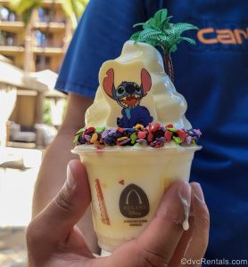 A hand holds a cup with the Aulani logo on it, which is filled with a Dole Whip Waffle Bowl topped with a white chocolate piece with Stitch on it, multicolored Mickey sprinkles, and a mini plastic palm tree.